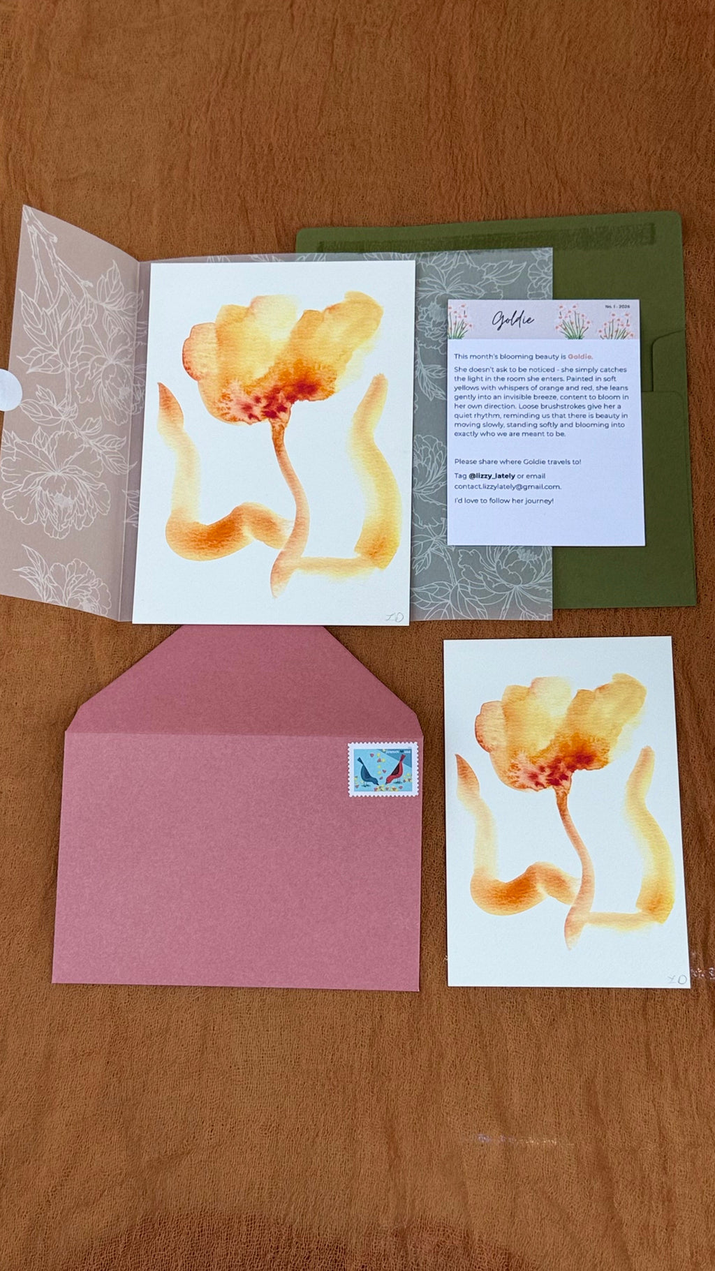 A floral stationery set arranged on a wooden surface, including an open white card with a yellow-orange flower illustration, a green envelope, a pink envelope, a smaller matching card, and a printed insert.